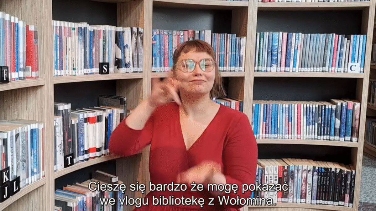 Na zdjęciu widać osobę w czerwonej bluzce, stojącą na tle regałów pełnych książek w bibliotece. Osoba wykonuje gesty rękami, prawdopodobnie w języku migowym. W tle widoczne są poukładane książki, a każdy regał oznaczony jest literami pomagającymi w orientacji. Na dole zdjęcia znajduje się napis w języku polskim: „Cieszę się bardzo że mogę pokazać we vlogu bibliotekę z Wołomina.”