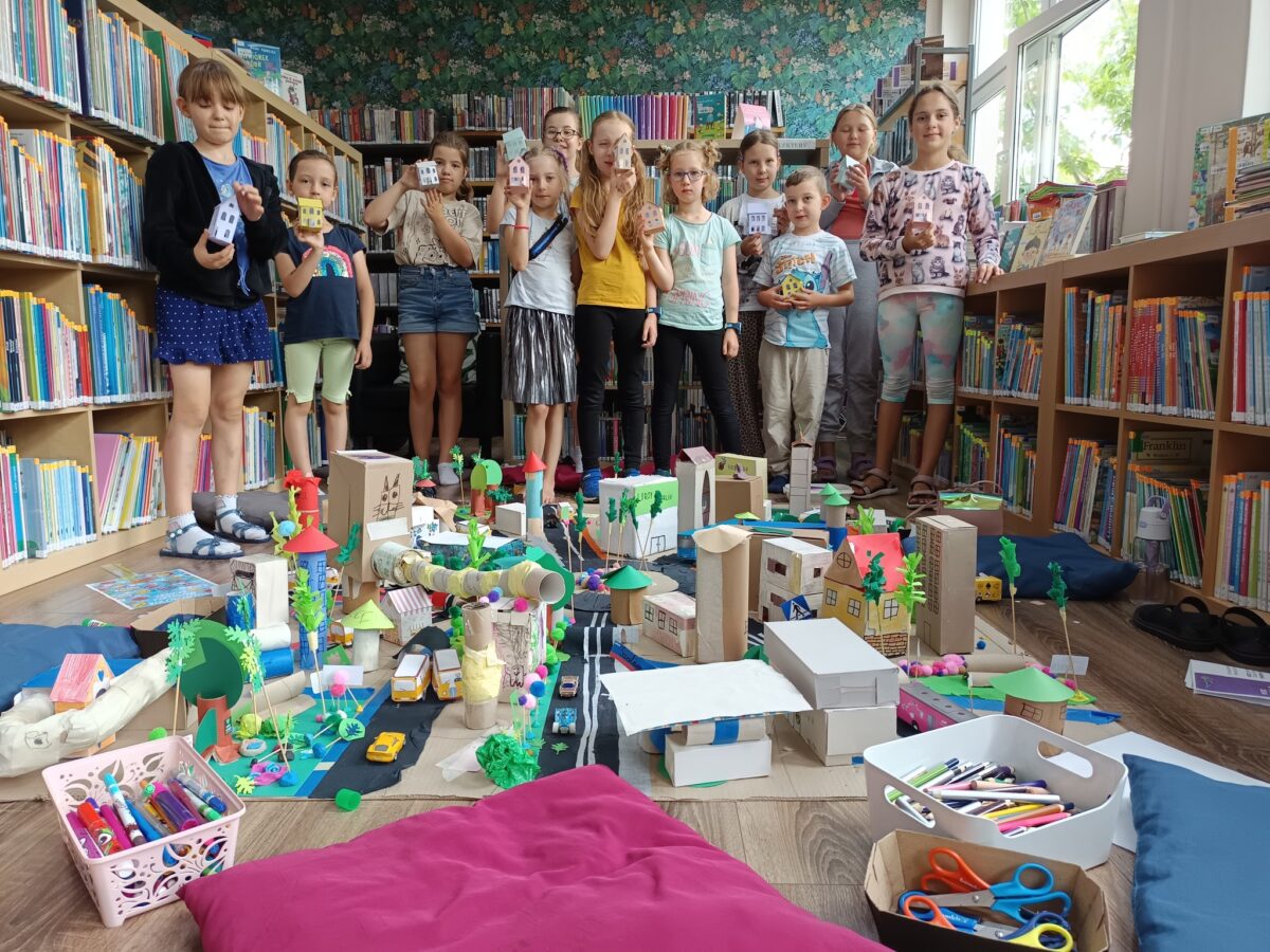 Na podłodze w kolorowej bibliotece stworzone zostało duże, przestrzenne miasto z różnorodnych materiałów plastycznych, takich jak kartonowe pudełka, rolki po papierze, papier kolorowy, patyczki, plastikowe nakrętki i pompony. Konstrukcja zawiera budynki, pojazdy, drzewa i drogi, wszystko urozmaicone barwnymi detalami i ręcznie wykonaną zielenią. Po obu stronach znajdują się wygodne poduszki oraz koszyki z przyborami do rysowania i wycinania, co podkreśla atmosferę twórczej zabawy. W tle widoczne są ściany pełne półek, które szczelnie wypełniają książki dziecięce, a z prawej strony wpada do pomieszczenia naturalne światło z dużego okna. Przy makiecie stoi grupa dzieci prezentujących własnoręcznie wykonane budowle, co nadaje scenie radosny i kreatywny wyraz.