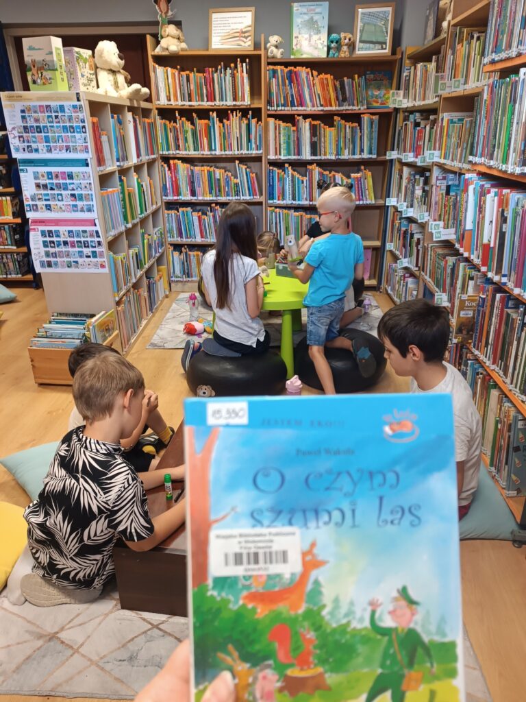  Zbliżenie na książkę pt. „O czym szumi las”, którą trzyma dorosła osoba. Tytuł jest napisany dużą czcionką w&nbsp;centralnej części okładki. W&nbsp;tle widać grupę dzieci, które siedzą na podłodze i&nbsp;przy małym stoliku, otoczone regałami z&nbsp;książkami, biorąc udział w&nbsp;kreatywnym warsztacie.