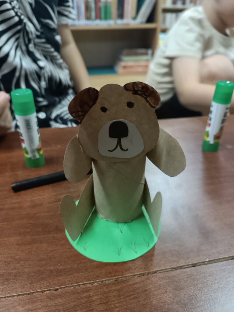 Zbliżenie na małą figurkę psa/misia, wykonaną z&nbsp;papierowej rolki w&nbsp;brązowym kolorze. Ma uszy, głowę i&nbsp;łapy, a&nbsp;w środku pyszczka ma białe kółko z&nbsp;namalowanymi oczami i&nbsp;nosem. Figurka stoi na zielonym okręgu z&nbsp;wyciętymi wzorami przypominającymi źdźbła trawy. W&nbsp;tle widać dłonie dwóch innych osób i&nbsp;kleje w&nbsp;sztyfcie.