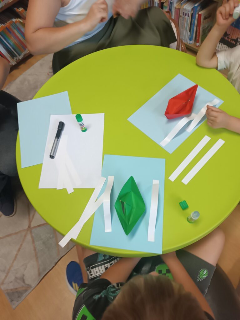 Ujęcie z&nbsp;góry na okrągły, zielony stół. Przy stole siedzi kilka osób. Na stole leżą materiały do rękodzieła, w&nbsp;tym niebieskie i&nbsp;białe kartki papieru, paski białego papieru, pisak i&nbsp;klej. W&nbsp;centrum stołu znajdują się dwie gotowe, papierowe łódki: jedna zielona, a&nbsp;druga czerwona.