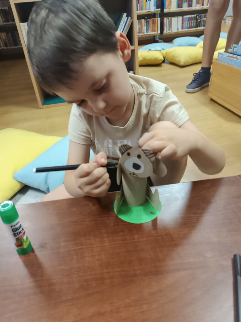 Zbliżenie na dziecko, które siedzi na podłodze i&nbsp;pracuje nad projektem plastycznym. Dziecko trzyma czarny pisak i&nbsp;rysuje na małej, brązowej figurce psa/misia, zrobionej z&nbsp;papierowej rolki. Figurka stoi na zielonym, okrągłym kawałku papieru, imitującym trawę.