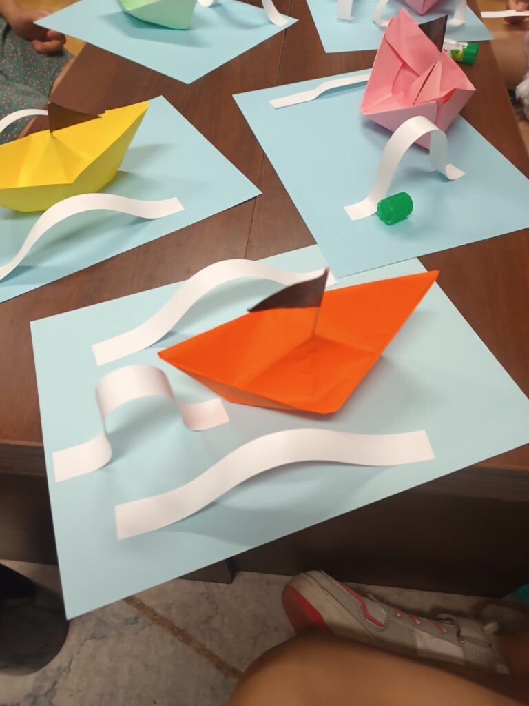  Zbliżenie na drewniany stół, na którym leżą papierowe łódki w&nbsp;kolorach żółtym, różowym i&nbsp;pomarańczowym. Łódki są ustawione na jasnoniebieskich arkuszach papieru, z&nbsp;każdej strony otoczone pofalowanymi, białymi paskami, które imitują fale. W&nbsp;rogu zdjęcia widać zielony klej w&nbsp;sztyfcie.