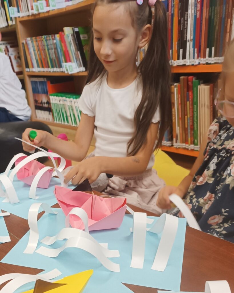 Zbliżenie na dziewczynkę z&nbsp;długimi, brązowymi włosami upiętymi w&nbsp;dwa kucyki, która siedzi przy stole i&nbsp;pracuje nad papierowymi łódkami. Dziewczynka trzyma zielony klej w&nbsp;sztyfcie i&nbsp;przykleja białe paski papieru, imitujące fale, do różowej łódki. Na stole widać inne, podobne prace.