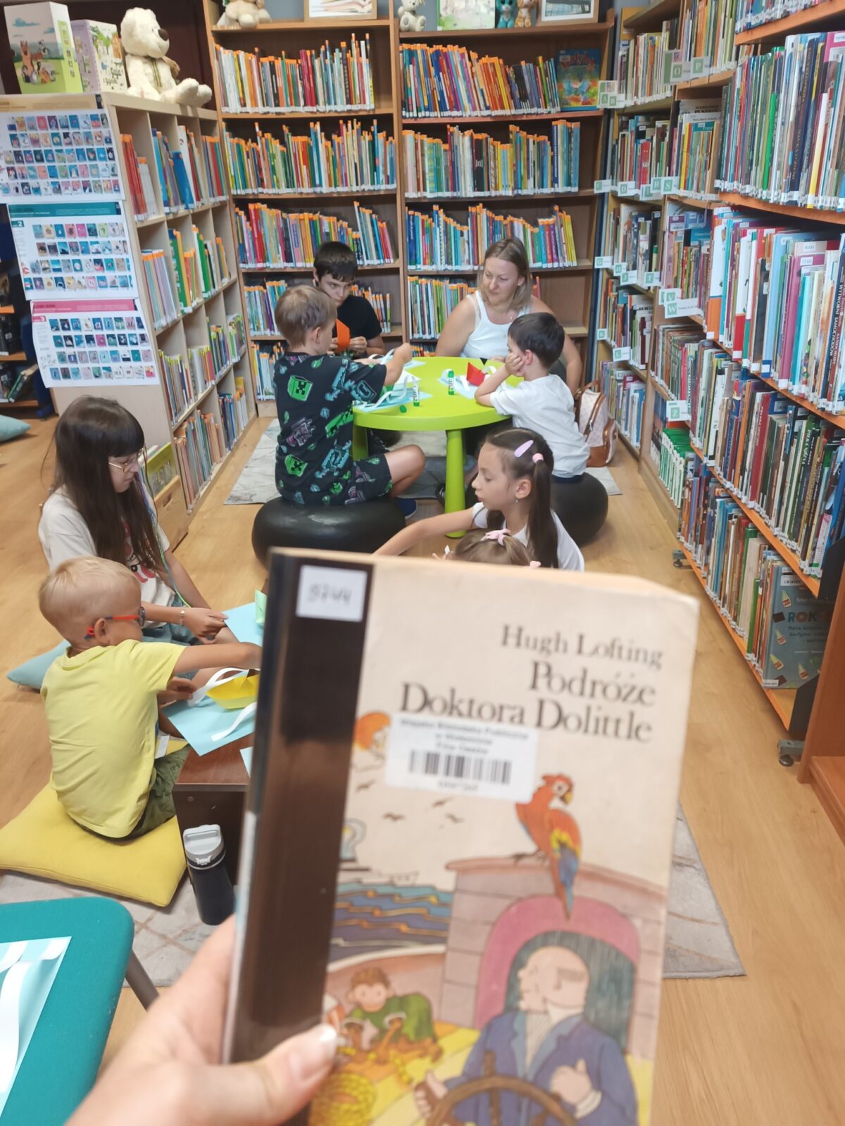 Na zdjęciu widać wnętrze kolorowej biblioteki dziecięcej, wypełnionej regałami z książkami. W centrum pomieszczenia stoi okrągły, limonkowy stolik, przy którym dzieci siedzą na kolorowych poduszkach oraz czarnych pufach. Zajmują się pracami plastycznymi, wspólnie wycinając i sklejając papier. Na regałach wokół nich znajdują się liczne książki – ich grzbiety są widoczne i ustawione w równych rzędach. Na pierwszym planie znajduje się ręka trzymająca książkę Hugh Loftinga „Podróże doktora Dolittle”; okładka jest w jasnych kolorach, ilustrowana rysunkiem przedstawiającym postać doktora oraz papugę siedzącą na rurze statku. Pomieszczenie jest przyjazne i pełne życia, idealnie stworzone do czytania i nauki, a na górnych półkach stoją pluszowe misie. Cała przestrzeń emanuje ciepłem oraz zachęca do wspólnego spędzania czasu w otoczeniu literatury dziecięcej.