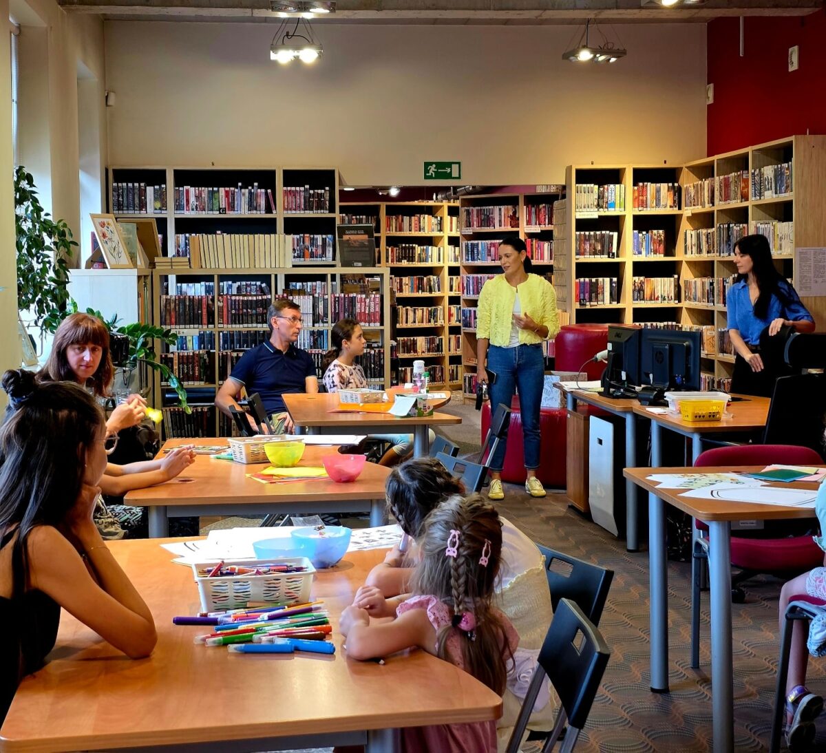 Na zdjęciu widoczna jest sala biblioteczna z jasnymi ścianami i wysokim sufitem. W tle znajdują się duże, drewniane regały wypełnione książkami ustawionymi w rzędach. Przy kilku ustawionych w pomieszczeniu stołach siedzą dzieci i dorośli, wspólnie uczestnicząc w zajęciach plastycznych – na stołach leżą kolorowe flamastry, kartki papieru, zestawy do rysowania, a także miski i koszyki na przybory. W centralnej części sali stoi osoba ubrana w żółty sweter i jasne spodnie, a obok niej dwie osoby prowadzące zajęcia – jedna z nich ubrana jest w niebieską koszulę. W pomieszczeniu panuje przyjazna, aktywna atmosfera sprzyjająca nauce i twórczości. Całość wnętrza jest dobrze doświetlona dzięki dużym oknom po lewej stronie, przez które wpada naturalne światło. W tle widać również rośliny doniczkowe oraz sprzęt komputerowy ustawiony na jednym ze stołów.