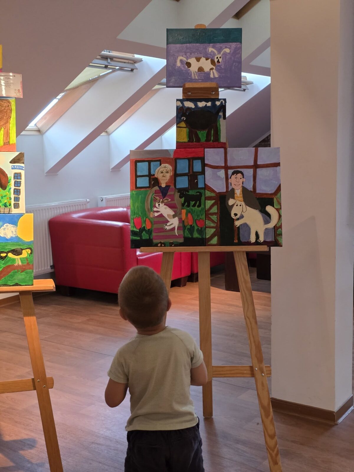 Na zdjęciu widać dziecko stojące tyłem, w jasnej koszulce i ciemnych spodenkach, które z zaciekawieniem ogląda obrazy umieszczone na sztalugach w jasnej, przestronnej galerii. Centralnie znajduje się sztaluga z kilkoma kolorowymi pracami malarskimi, przedstawiającymi różne sceny ze zwierzętami domowymi: na najwyższym obrazie namalowany jest pies w kremowo-brązowe łaty na fioletowym tle, poniżej widać czarnego kota na zielonej trawie, a na dolnych dwóch obrazach – sceny w gospodarstwie, gdzie wśród kwiatów białe i czarne koty bawią się z dzieckiem, a dorosła postać trzyma na smyczy dużego białego psa. W tle widoczny jest czerwony fotel, ukośnie ustawione okna dachowe, jasne ściany oraz ciepła, drewniana podłoga, które nadają miejscu przytulny klimat. Cała scena jest pogodna i spokojna, a motyw przewodni obrazów skupia się na przyjaźni człowieka ze zwierzętami.
