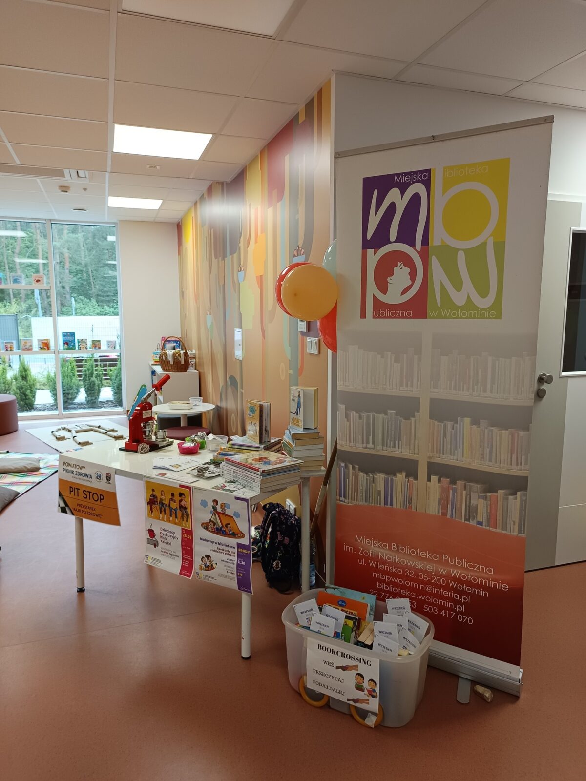 Na zdjęciu znajduje się jasne, kolorowo urządzone wnętrze pomieszczenia, przypominające przestrzeń przeznaczoną dla dzieci. Po lewej stronie widoczna jest duża witryna okienna, przez którą widać zieleń na zewnątrz. Przy ścianie po prawej stronie stoi rollup z kolorowym logotypem Miejskiej Biblioteki Publicznej w Wołominie oraz z informacjami kontaktowymi tej instytucji. Przed rollupem stoi plastikowy pojemnik z napisem "BOOKCROSSING - weź, przeczytaj, podaj dalej", w którym znajdują się książki i broszury. Obok pojemnika ustawiono biały stolik, na którym znajduje się stosik książek, zabawki, ulotki oraz plakaty informacyjne. Na ścianie i przy stoisku wiszą balony w kolorach czerwonym, żółtym i zielonym. W głębi pomieszczenia widać miękkie maty i wyłożone zabawki, które nadają przestrzeni przyjazny i zachęcający do zabawy charakter. Całość tworzy wrażenie miejsca sprzyjającego rodzinnej, edukacyjnej aktywności oraz promującego czytelnictwo i wymianę książek.