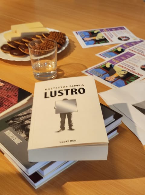 Zdjęcie przedstawia drewniany stół, na którym leży kilka przedmiotów. Na pierwszym planie znajduje się książka Krzysztofa Blińki o tytule „Lustro", ułożona na kilku innych książkach. Okładka ukazuje sylwetkę osoby w szarych spodniach, trzymającej przed sobą prostokątne lustro, w którym odbija się tylko jasne tło. Po lewej stronie stoi szklanka z przezroczystą wodą, a nieco dalej, z tyłu po lewej, znajduje się biały talerz z różnymi ciastkami oraz prostokątnymi kawałkami sera. W prawym górnym rogu leżą wydrukowane kolorowe ulotki – są wyraźnie widoczne ilustracje twarzy oraz napisy, choć szczegóły tekstu są nieczytelne. Po prawej stronie zdjęcia widać też białe kartki, częściowo nachodzące na stos książek. Całość sugeruje kameralne spotkanie przy stole lub przygotowania do czytania i rozmowy przy przekąskach.