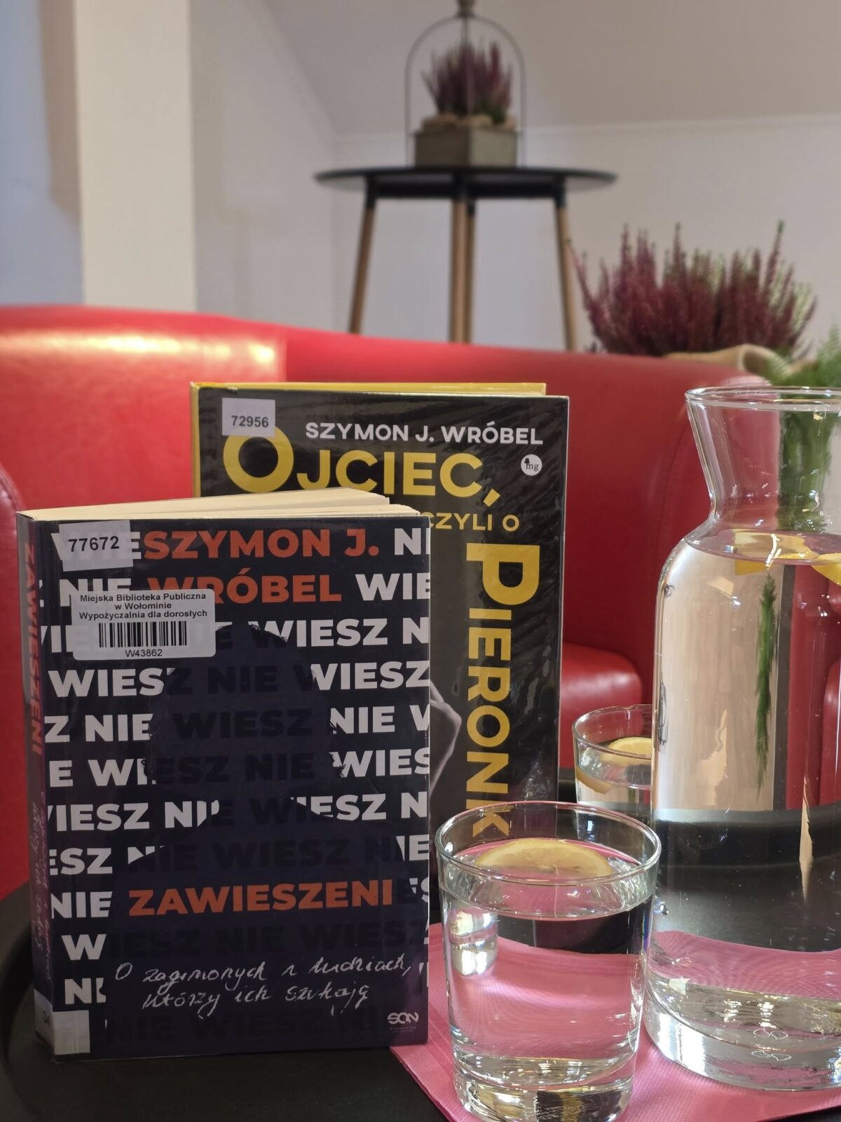 Na pierwszym planie znajduje się okrągły czarny stolik, na którym ustawiono dwie książki i szklany dzbanek z wodą oraz plasterkiem cytryny. Obok dzbanka stoją dwie szklanki, w jednej z nich również znajduje się plasterek cytryny. Książki ustawione są pionowo i lekko na siebie nachodzą — ich okładki są dobrze widoczne. Obie pozycje są autorstwa Szymona J. Wróbla, a tytuły to: „Ojciec, czyli o Pieronku” oraz „Nie wiesz, wiesz? Zawieszeni”. W tle widać fragment czerwonej sofy, wysokie białe ściany oraz osobny stolik z ozdobną doniczką i wrzosami. Całość sprawia wrażenie przytulnego, jasnego wnętrza o kameralnej atmosferze.