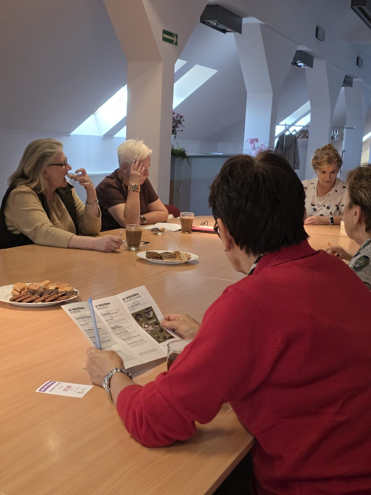 Zdjęcie przedstawia grupę sześciu starszych osób siedzących przy dużym, jasnym, drewnianym stole w dobrze oświetlonym pomieszczeniu o nowoczesnym wystroju. Na stole stoją filiżanki z kawą oraz dwa talerze zawierające ciastka i wafle. Jedna z kobiet, ubrana w czerwoną bluzę, przegląda ulotkę lub program spotkania, podczas gdy pozostałe osoby prowadzą rozmowę i wypoczywają. W tle widoczne są okna dachowe wpuszczające naturalne światło oraz kwiaty doniczkowe i wieszak na ubrania. Atmosfera miejsca wydaje się spokojna i sprzyja rozmowom towarzyskim.