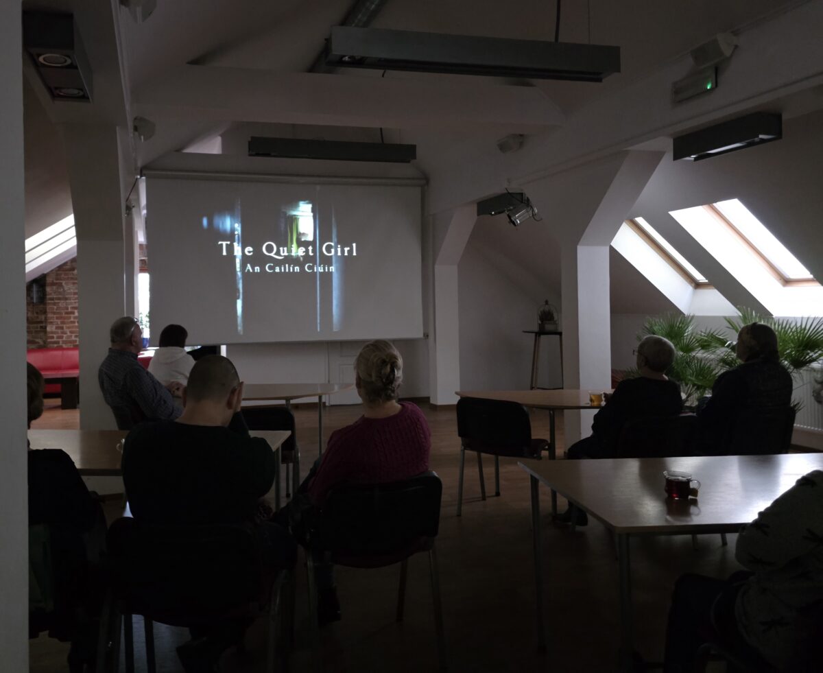 W przestronnej, jasnej sali znajdującej się na poddaszu kilkoro ludzi siedzi przy rozstawionych stołach, oglądając projekcję filmu. Pomieszczenie ma skośne sufity i duże połaciowe okna, przez które wpada naturalne światło. Ściany są białe, a część jednej z nich wykończona jest czerwoną cegłą. Ekran projekcyjny ustawiony z przodu sali wyświetla tytuł filmu: „The Quiet Girl – An Cailín Ciúin” w białych literach na ciemnym tle. Widzowie siedzą na krzesłach w niewielkich grupach; kilka osób zajmuje miejsca bliżej ekranu, a inni rozproszeni są przy stołach po bokach. Wśród nich są osoby o różnych sylwetkach i w różnym wieku, widziane głównie od tyłu lub z profilu. Niektórzy lekko pochylają głowy, wpatrzeni w ekran. Na stołach stoją szklanki z ciemnym napojem, być może herbatą. W lewym rogu sali widać czerwoną kanapę, a w prawym – kilka dużych roślin doniczkowych. Nad głowami widzów zawieszone są projektory oraz lampy sufitowe. Całość sprawia wrażenie kameralnego, spokojnego pokazu filmowego zorganizowanego w jasnej, przyjaznej przestrzeni.