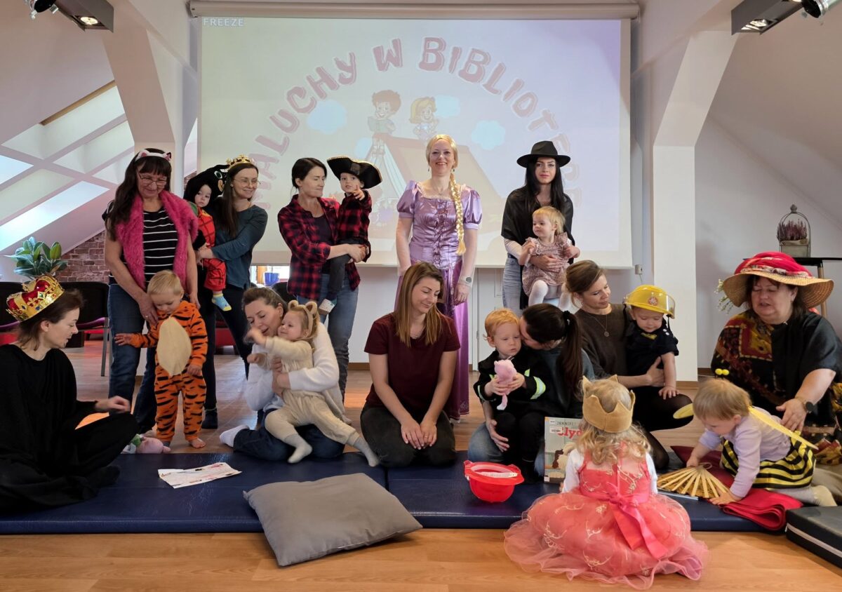 Na zdjęciu widoczna jest grupa dzieci oraz dorosłych, zgromadzona w jasnym, przestronnym pomieszczeniu z drewnianą podłogą. W tle, na ekranie, widać rysunkowy napis „Maluchy w bibliotece” oraz dwójkę rysunkowych dzieci. Większość osób jest przebrana w kolorowe stroje – są tutaj dzieci ubrane między innymi za tygryska, lwa, pszczółkę, księżniczkę w koronie oraz strażaka; dorośli mają na sobie m.in. kapelusze, peleryny i kostiumy nawiązujące do bajek lub baśni. Jedna osoba stoi na środku w stroju długowłosej księżniczki w fioletowej sukni. Przed grupą, na niebieskich matach do siedzenia, rozłożone są poduszki oraz kilka zabawek i książek. Panuje radosna, bajkowa atmosfera typowa dla czytania bajek lub zabawy tematycznej w bibliotece.