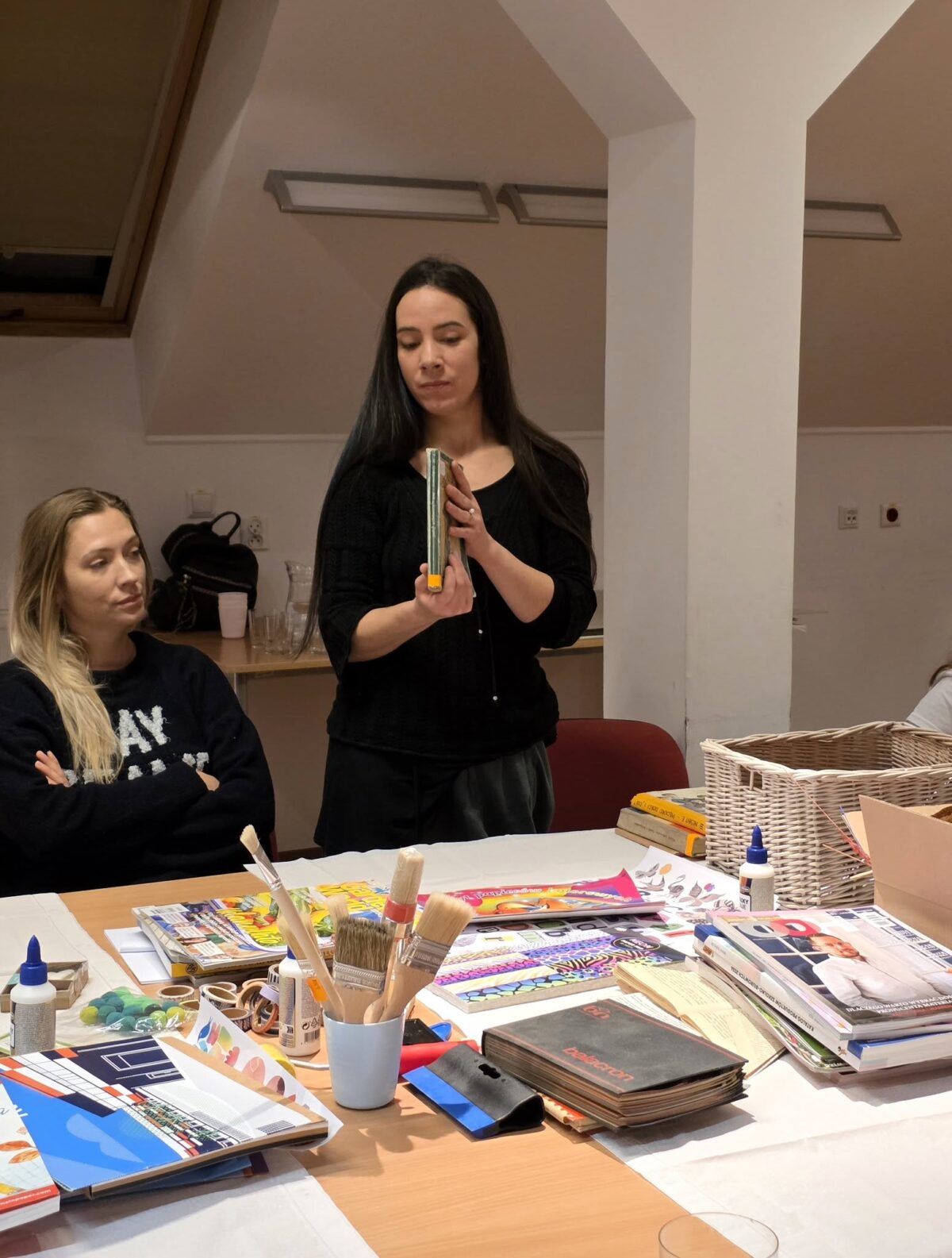 W jasnym pomieszczeniu przy dużym stole siedzi kilka osób, a na pierwszym planie widać rozłożone materiały plastyczne: pędzle w kubku, kleje, kolorowe czasopisma, kartony, taśmy i inne przybory do prac ręcznych. W centrum zdjęcia stoi kobieta o długich, ciemnych włosach, ubrana na czarno, która trzyma w dłoniach zamknięty notes lub książkę i pokazuje ją pozostałym, jakby tłumaczyła jakiś etap pracy. Po lewej stronie siedzi młoda kobieta z jasnymi włosami, z założonymi rękami, uważnie przyglądająca się prowadzącej. W tle widać białe ściany, skośny sufit i kosz wiklinowy na końcu stołu, co nadaje scenie charakteru warsztatów artystycznych lub zajęć kreatywnych.
