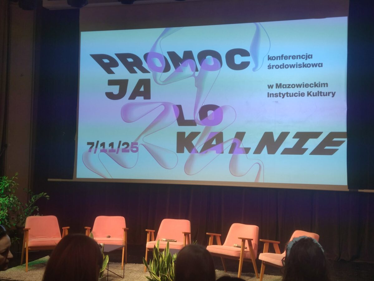 Na zdjęciu widać scenę przygotowaną do konferencji w sali teatralnej lub kinowej. Na tle ciemnej kurtyny wisi duży ekran z kolorową grafiką i napisem po polsku „PROMOCJA LOKALNIE”, obok którego znajdują się mniejsze napisy „konferencja środowiskowa w Mazowieckim Instytucie Kultury” oraz data „7/11/25”. Przed ekranem ustawiono w jednym rzędzie pięć różowych foteli na drewnianych nogach, pomiędzy którymi stoją małe stoliki z mikrofonami; z boków sceny oraz na przodzie znajdują się donice z zielonymi roślinami. Na dolnej krawędzi zdjęcia widać zarys kilku osób siedzących na widowni, patrzących w stronę sceny.