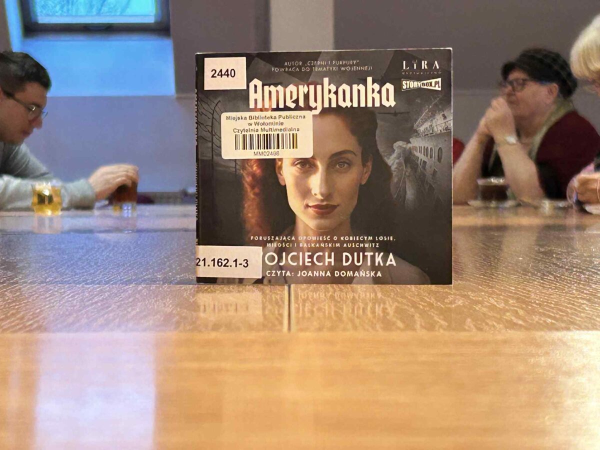 Na pierwszym planie stoi na stole pudełko audiobooka z tytułem „Amerykanka”, na którym widać fragment twarzy kobiety i nadrukowane biblioteczne naklejki inwentarzowe. Za pudełkiem, przy długim drewnianym stole, siedzi kilka osób pijących napoje w szklankach, a całość odbywa się w jasnej sali z dużym oknem w tle.