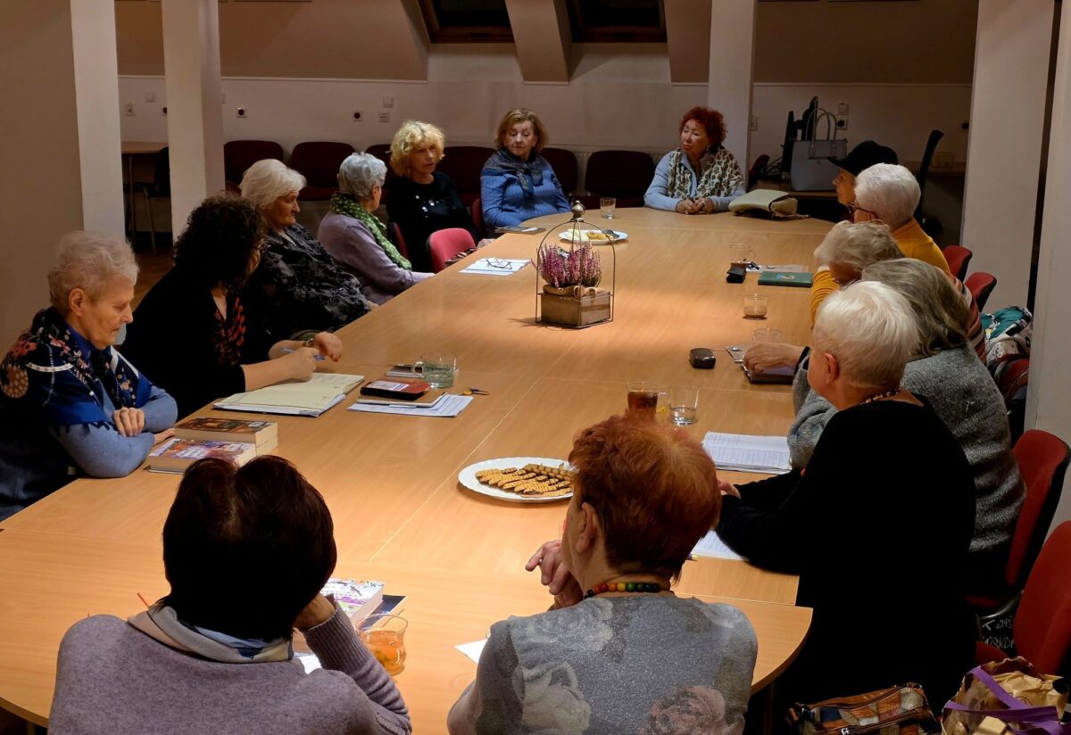 Na zdjęciu widać dużą, jasną salę z długim, prostokątnym stołem zajmującym prawie cały kadr. Wokół stołu siedzi kilkanaście starszych kobiet, ubrane są swobodnie, w kolorowe swetry, chusty i szale, większość ma siwe włosy. Wszystkie są zwrócone do środka stołu i wyglądają, jakby uczestniczyły w spokojnej, uważnej rozmowie lub spotkaniu klubu dyskusyjnego – część notuje w zeszytach, przed niektórymi leżą książki i dokumenty, a na blacie stoją szklanki z napojami. Na środku stołu znajduje się dekoracja z wrzosem w małej doniczce oraz talerze z ciasteczkami, co nadaje scenie kameralny, gościnny charakter. W tle widać ściany z oknami dachowymi i rząd pustych krzeseł, co sugeruje, że jest to sala w domu kultury lub bibliotece.