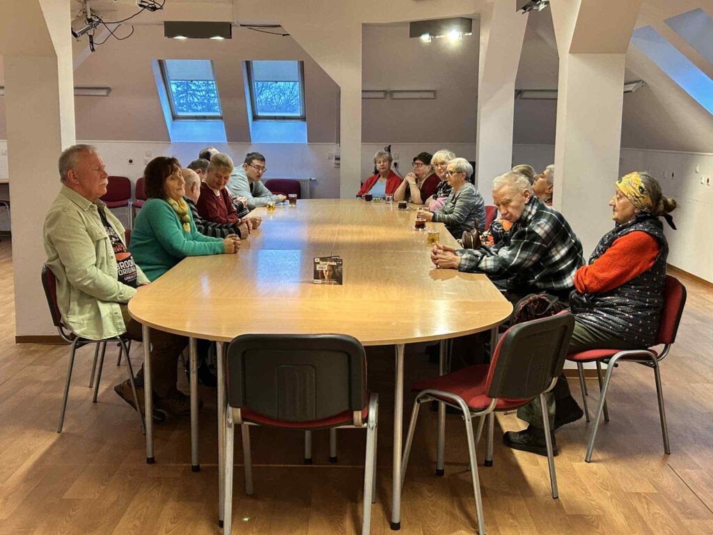 Fotografia pokazuje tę samą salę z&nbsp;innej perspektywy: kilkanaście osób siedzi wokół podłużnego, zaokrąglonego stołu, zwróconych w&nbsp;lewo, jakby słuchali prowadzącego spoza kadru. Na środku stołu stoi małe pudełko audiobooka, a&nbsp;za uczestnikami widać skośne okna dachowe i&nbsp;jasne ściany poddasza.