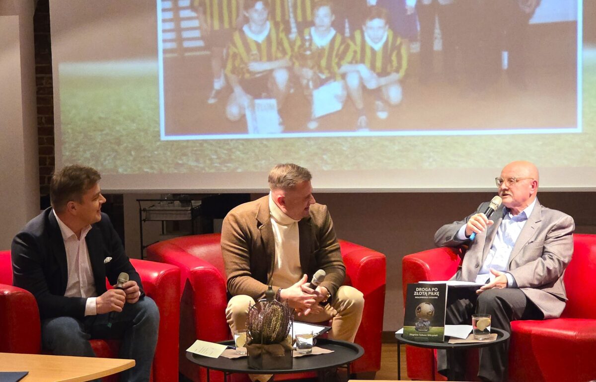 Na zdjęciu widać trzyosobową rozmowę na żywo w sali zaaranżowanej jak małe studio. Trzej mężczyźni siedzą w półokręgu na czerwonych, klubowych fotelach i trzymają mikrofony, jak podczas spotkania autorskiego lub panelu dyskusyjnego. Po lewej siedzi mężczyzna w ciemnej marynarce i białej koszuli, pośrodku mężczyzna w brązowej marynarce i jasnym golfie, a po prawej mężczyzna w jasnoszarej marynarce. Przed nimi stoją dwa niskie, okrągłe stoliki – na jednym leży kilka kartek i stoi dekoracyjna roślina w doniczce, na drugim widać szklankę z napojem i książkę o piłce nożnej z tytułem „Droga po Złotą Piłkę”. W tle, na dużym ekranie, wyświetlone jest rozmyte zdjęcie drużyny piłkarskiej w żółto‑czarnych strojach, co sugeruje, że rozmowa dotyczy futbolu lub kariery sportowej. Całość odbywa się w ciepłym, przytulnym oświetleniu, w kameralnej przestrzeni z elementami ceglanej ściany po lewej.