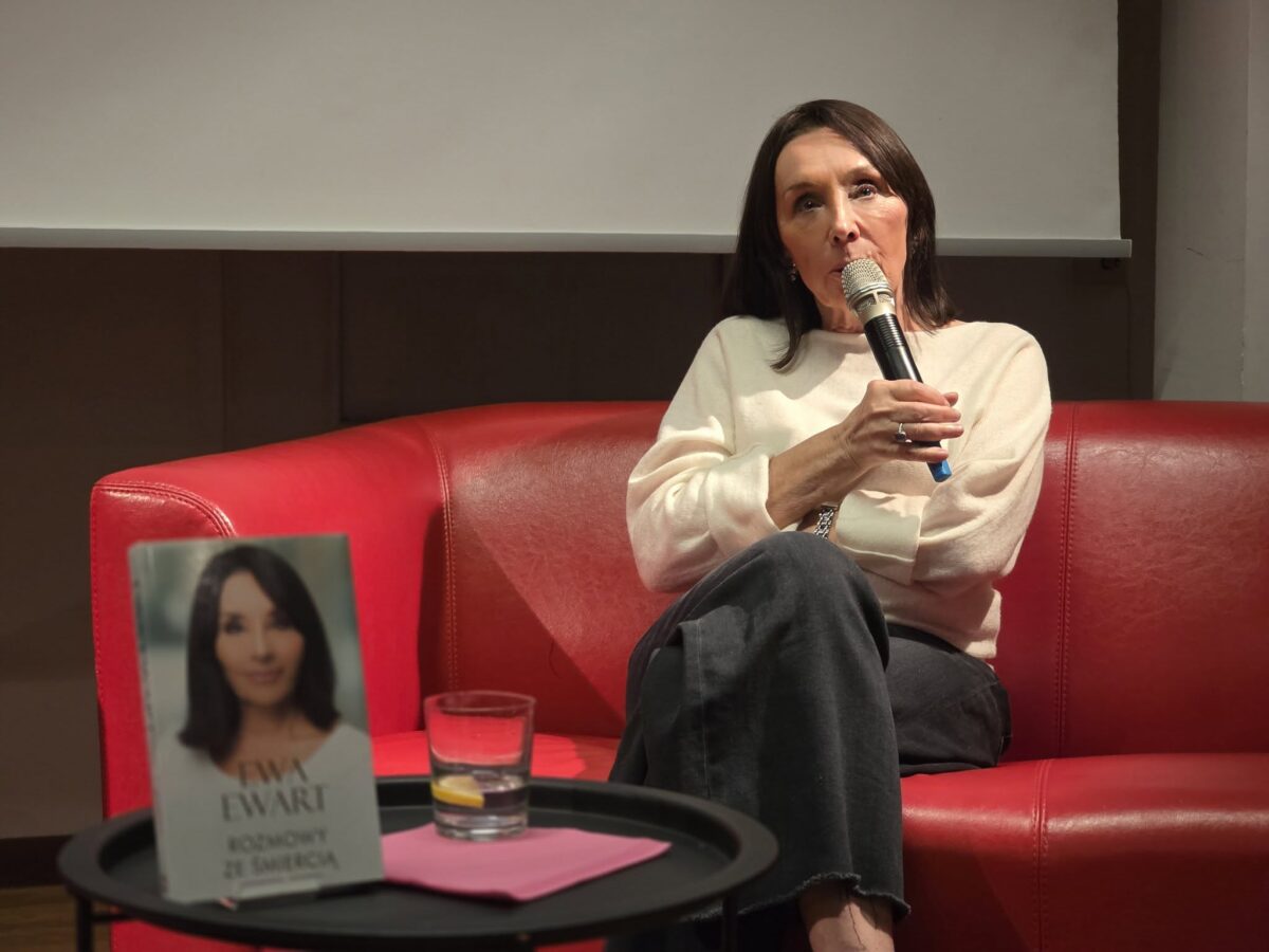 Na zdjęciu widać kobietę siedzącą na czerwonej, skórzanej kanapie. Ubrana jest w jasny sweter i ciemne spodnie. Trzyma mikrofon blisko ust, wygląda na skupioną i zaangażowaną w rozmowę. Przed nią, na czarnym stoliku, stoją szklanka z wodą oraz książka z jej portretem na okładce. Na książce widnieje napis: "Ewa Ewart - Rozmowy ze śmiercią". Tło jest stonowane, w górnej części widać biały ekran projekcyjny.