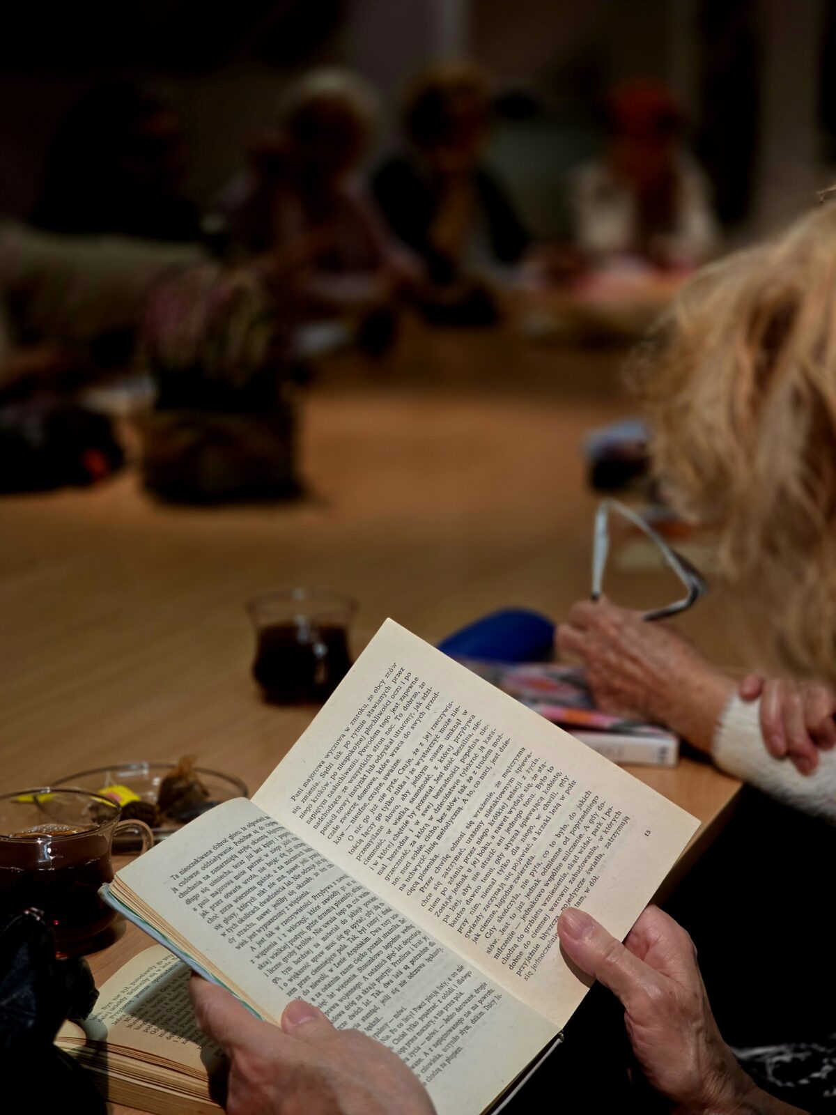 Zbliżenie na dłonie starszej osoby trzymającej otwartą książkę przy dużym stole. W tle, lekko rozmytym, siedzi kilka innych osób, również zajętych czytaniem lub rozmową. Na stole stoją kubki z herbatą oraz drobne przedmioty. Scena przypomina spotkanie klubu książki lub wspólne czytanie.