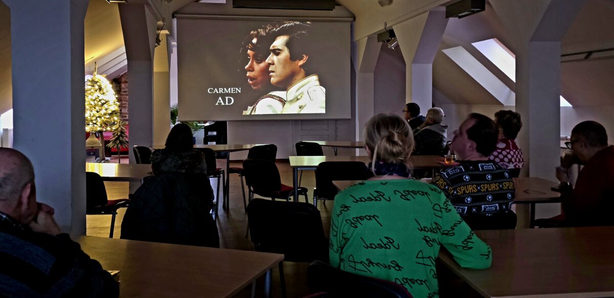 Uczestnicy spotkania siedzą przy stołach w bibliotecznej sali i oglądają projekcję opery „Carmen” wyświetlaną na dużym ekranie. Wnętrze jest jasne, z charakterystycznymi skosami poddasza, w tle widać świątecznie udekorowaną choinkę. Publiczność skupiona jest na seansie, panuje spokojna, kameralna atmosfera wspólnego odbioru kultury.