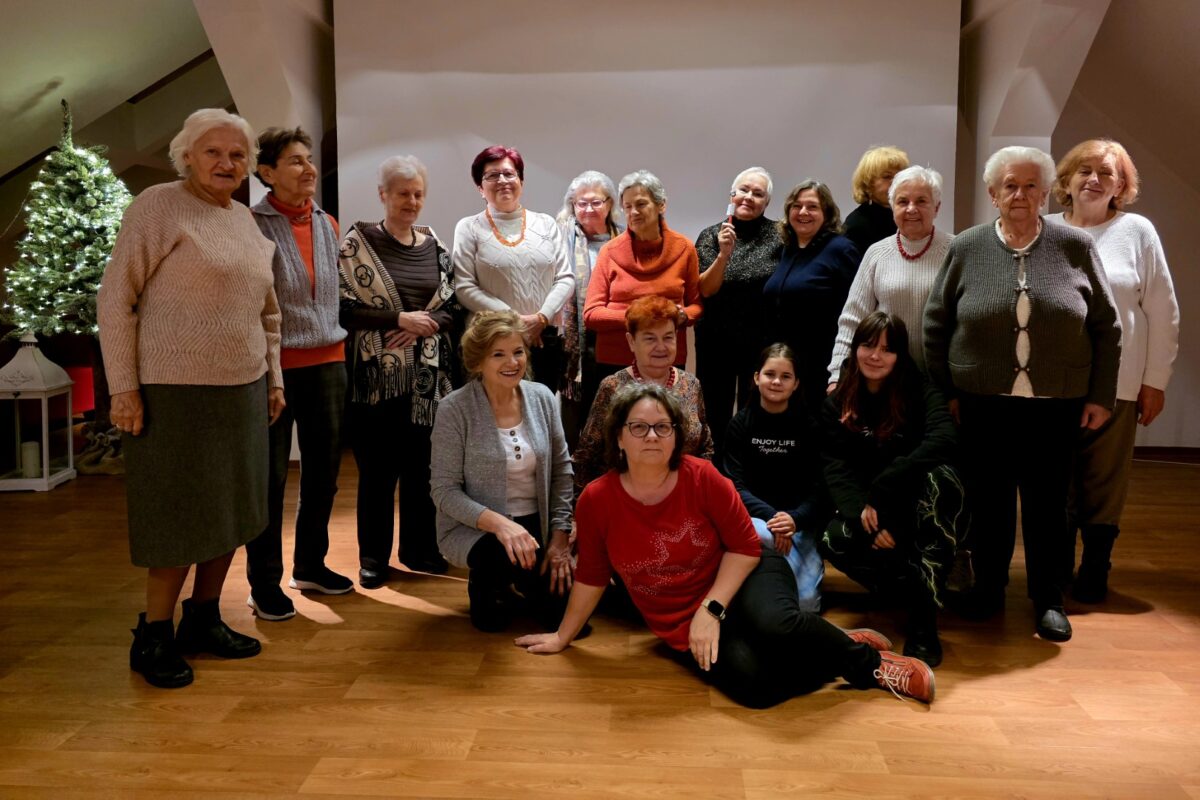 Grupowe zdjęcie kilkunastu kobiet w różnym wieku, od młodszych po seniorki, pozujących w sali o drewnianej podłodze. Część osób stoi, część klęczy lub siedzi z przodu. Uczestniczki ubrane są w ciepłe, codzienne stroje, swetry, bluzki, spodnie, w stonowanych kolorach z akcentami czerwieni i pomarańczu. Większość patrzy w stronę aparatu i uśmiecha się. Po lewej stronie widoczna jest udekorowana, oświetlona choinka, co sugeruje zimowy lub świąteczny czas spotkania. Atmosfera zdjęcia jest spokojna i serdeczna, przedstawia wspólnotę i międzypokoleniowe spotkanie.