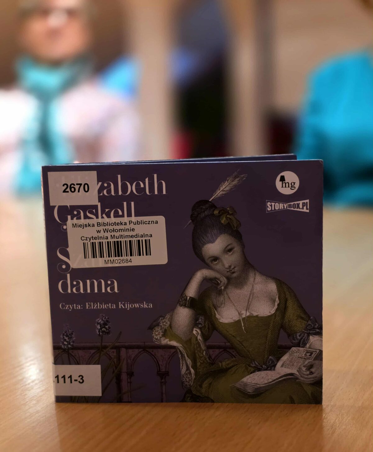 Na pierwszym planie stoi pudełko z audiobookiem autorstwa Elizabeth Gaskell. Okładka jest fioletowa, z ilustracją XIX-wiecznej kobiety w zielonej sukni, siedzącej i czytającej książkę. Widoczny jest tytuł zakończony słowem „dama”, informacja „Czyta: Elżbieta Kijowska” oraz logo Storybox.pl. Na okładce znajdują się także naklejki biblioteczne z numerem inwentarzowym. W tle, nieostre wnętrze biblioteki z siedzącymi osobami.