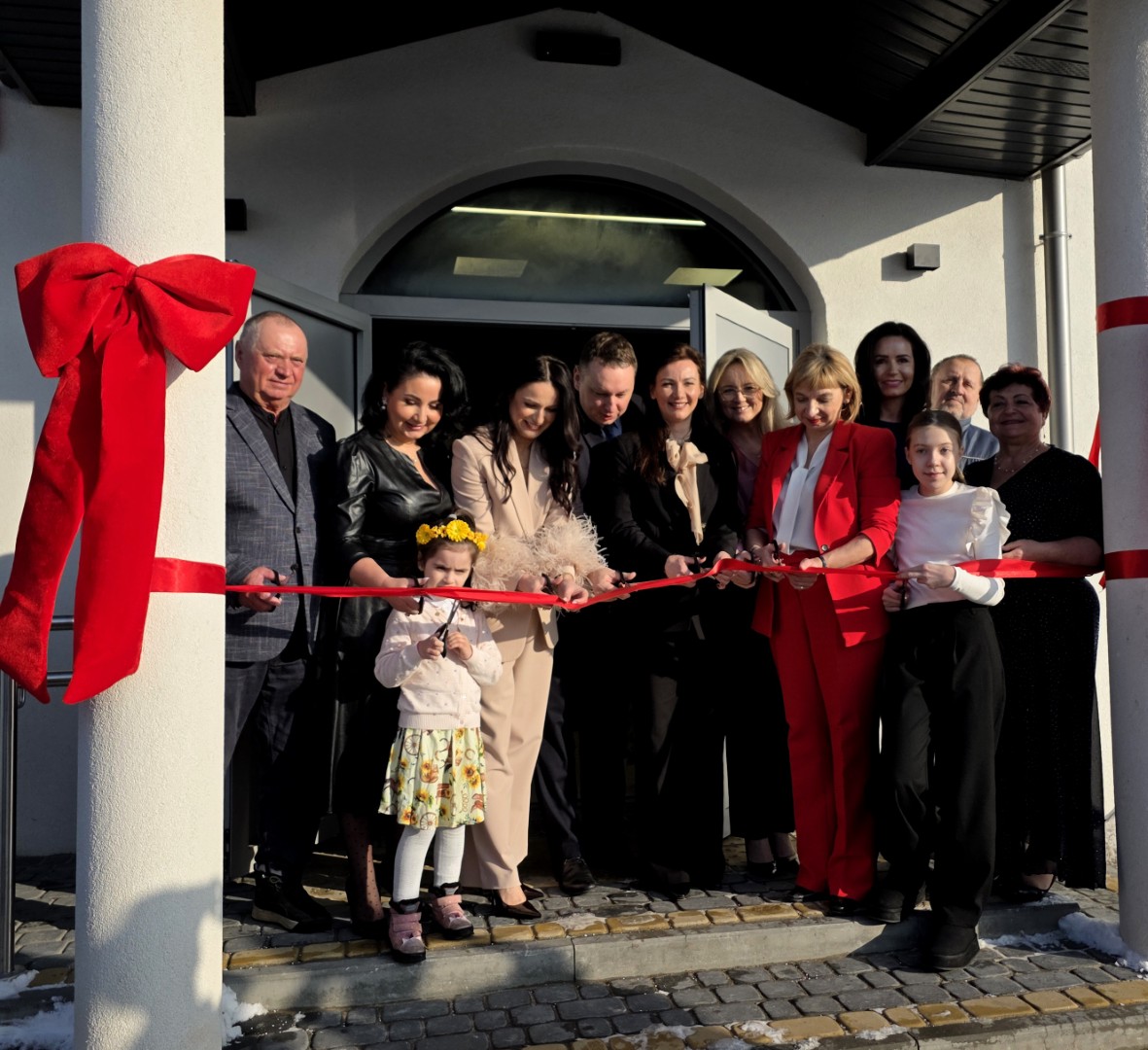 Grupa dorosłych i dzieci stoi przed wejściem do jasnego budynku z otwartymi drzwiami, tworząc rząd na schodku nad brukowanym chodnikiem; wszyscy trzymają rozpiętą, czerwoną wstęgę przeciągniętą między dwoma białymi filarami, z których lewy jest udekorowany dużą, czerwoną kokardą, co wskazuje na uroczyste otwarcie obiektu w słoneczny, zimowy dzień.
