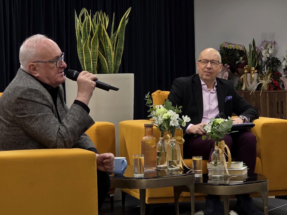 Dwóch mężczyzn siedzi naprzeciw siebie w żółtych fotelach podczas rozmowy na żywo w zamkniętym pomieszczeniu; po lewej stronie starszy mężczyzna w szarej marynarce trzyma w dłoni mikrofon, po prawej młodszy, elegancko ubrany w czarną marynarkę i jasną koszulę, trzyma notes lub tablet na kolanach. Pomiędzy nimi stoją dwa niskie stoliki z kilkoma szklanymi butelkami wody, szklankami oraz wazonikami z jasnymi kwiatami i zielonymi liśćmi, obok ułożono kilka książek. W tle widać ciemną zasłonę, wysoką donicę z długimi, pionowymi liśćmi oraz szafkę po prawej stronie, na której ustawiono bukiety kwiatów ozdobione wstążkami.