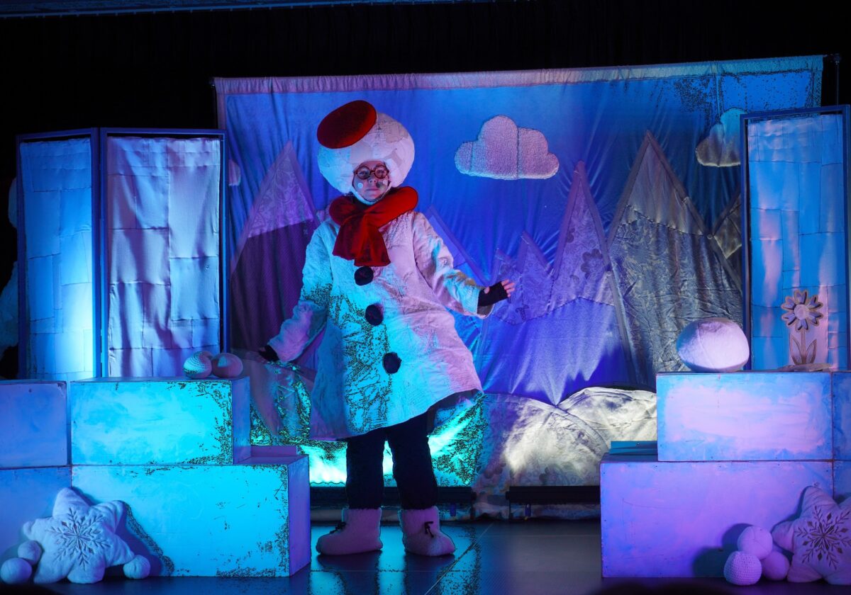 Sceniczna dekoracja zimowa: pośrodku stoi postać przebrana za bałwana w białym, lekko przedłużonym płaszczu z trzema dużymi czarnymi guzikami, czerwonym szalikiem i czerwonym kapeluszem przekrzywionym na bok, na tle błękitnej kurtyny z aplikacjami gór, chmur i śniegu; po obu stronach znajdują się białe prostopadłościenne „lodowe” bloki i pluszowe śnieżne dekoracje.