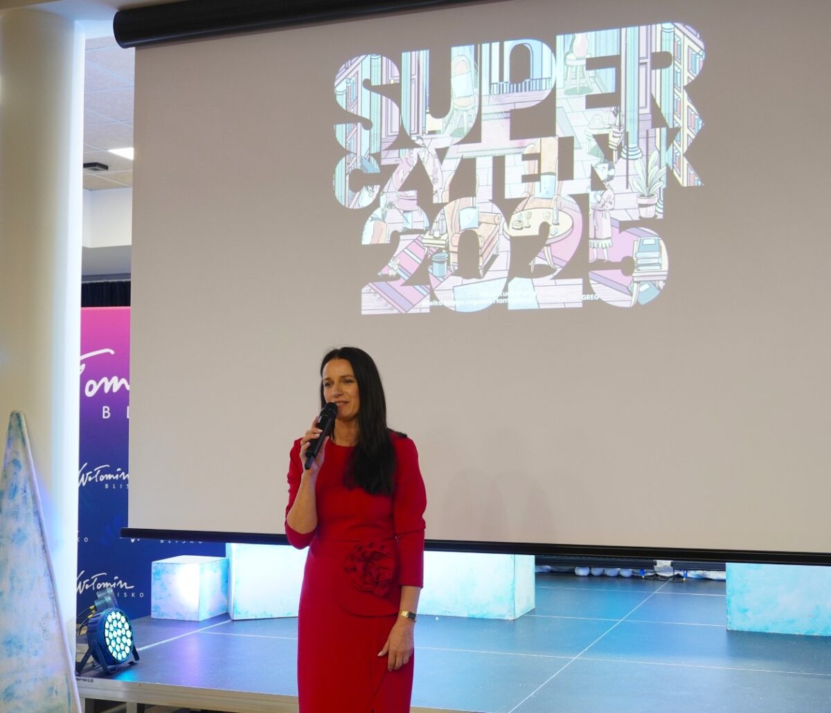 Kobieta w długiej, dopasowanej czerwonej sukience stoi na scenie z mikrofonem w dłoni, zwrócona przodem do publiczności. Za nią wisi duży biały ekran z kolorowym napisem „SUPERCZYTELNIK 2025”, wypełnionym rysunkową ilustracją wnętrza biblioteki. Scena jest jasno oświetlona, a po bokach ekranu widać podświetlane na niebiesko prostopadłościenne elementy dekoracyjne oraz fragment stojącego banera z napisem „Wołomin blisko”.