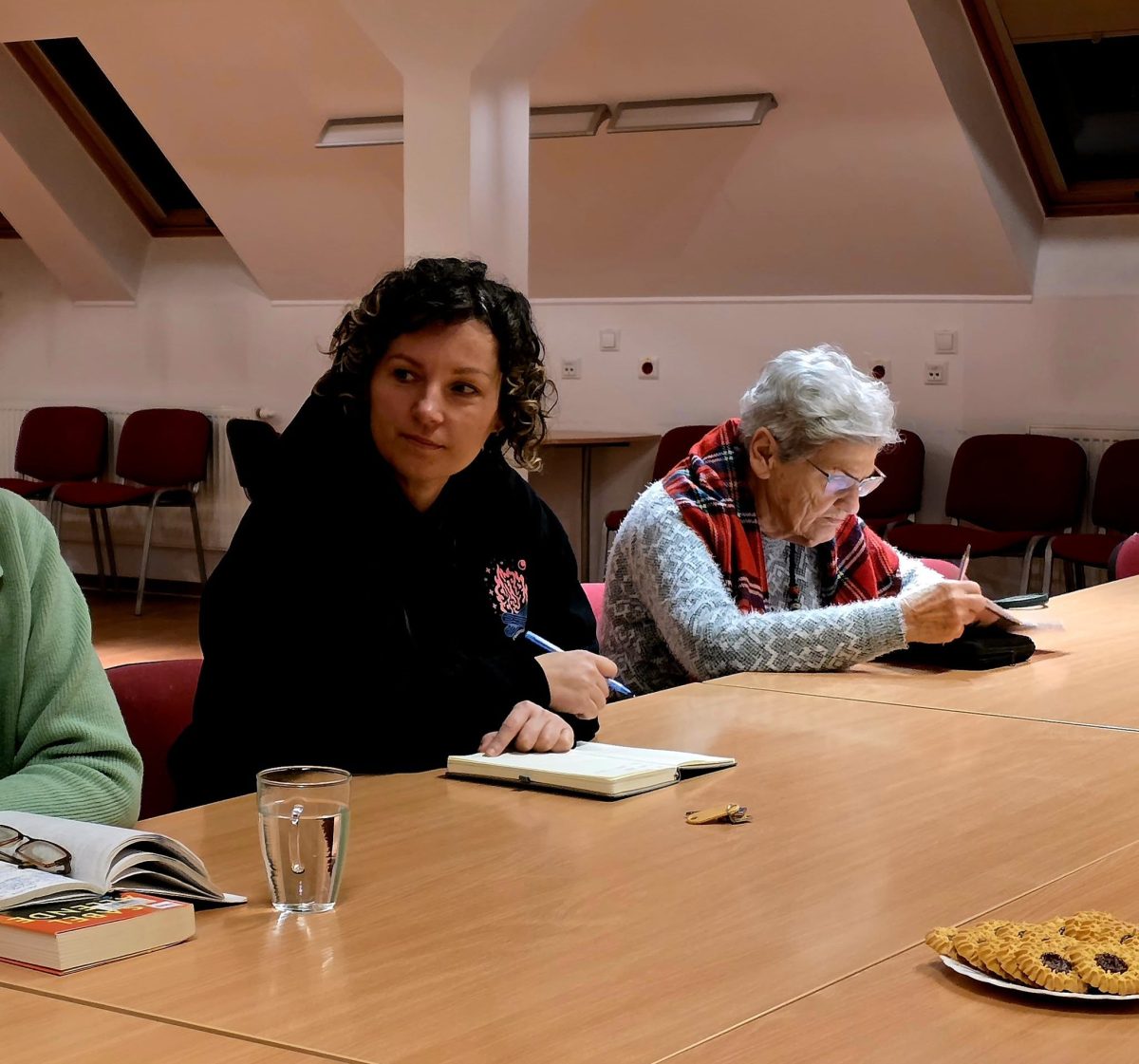 Kilka osób siedzi przy dużym, jasnobrązowym stole w sali spotkań, przypominającej świetlicę lub salę biblioteczną. Na pierwszym planie widać fragment otwartej książki, okulary i szklankę wody postawione na stole. W centrum siedzi młodsza osoba w ciemnej bluzie, przed sobą ma otwarty notes i trzyma długopis, jakby robiła notatki. Obok niej siedzi starsza osoba z siwymi włosami, w swetrze i kraciastym szalu, zapisująca coś w notatniku leżącym na stole. W tle stoją rzędy pustych, czerwonych krzeseł pod skośnym sufitem z oknami dachowymi, a z prawej strony na stole leży talerz z ciastkami.