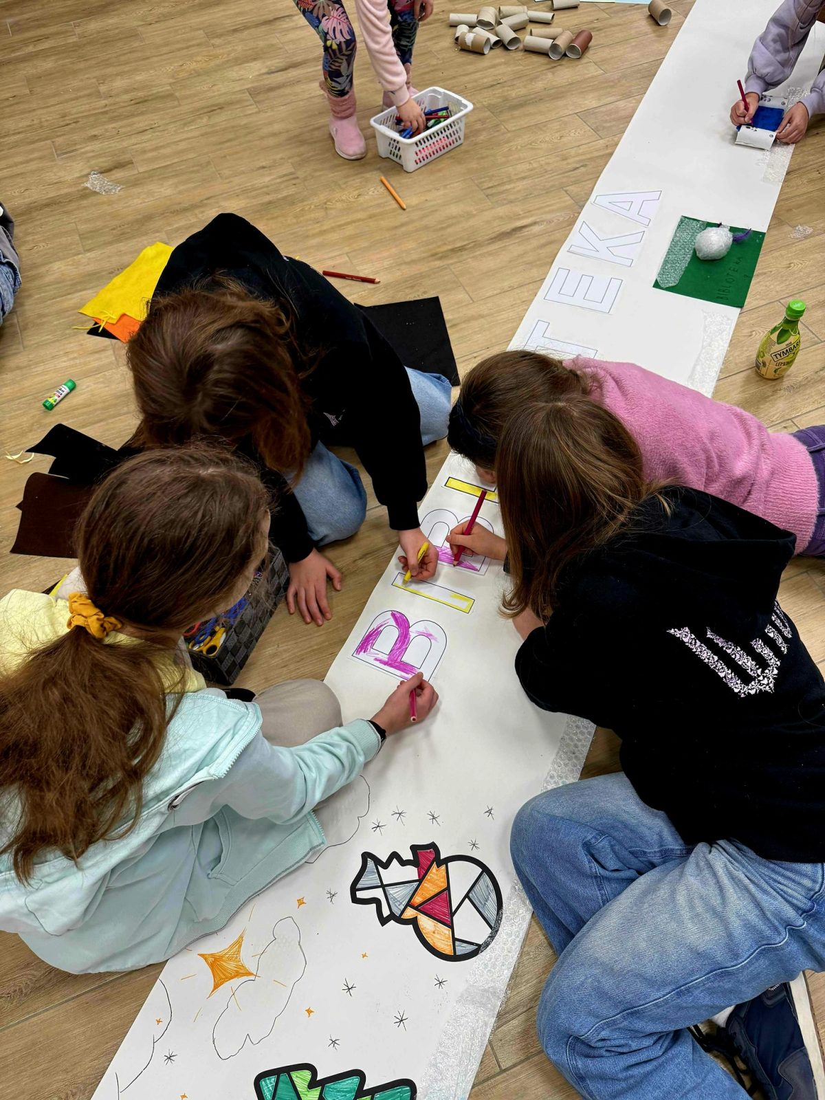 Kilkoro dzieci siedzi na drewnianej podłodze i wspólnie ozdabia długi, biały papierowy baner rozłożony wzdłuż sali. Na środku widoczne są duże kontury liter kolorowanych kredkami, z których część układa się w napis „BIBLIOTEKA”. W dolnej części baneru narysowane są proste kolorowe ilustracje, między innymi choinka i bombka o geometrycznym wzorze. Dzieci trzymają kredki i klej, obok leży koszyk z przyborami plastycznymi oraz kartonowe rolki i kawałki kolorowych tkanin. Cała scena przedstawia zajęcia plastyczne w pomieszczeniu o jasnej, gładkiej podłodze.