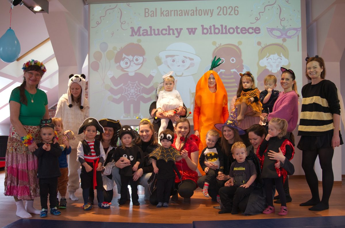 Grupa dorosłych i kilkunastu małych dzieci pozuje we wnętrzu biblioteki podczas balu karnawałowego, wszyscy mają kolorowe przebrania takie jak marchewka, pszczoła, pirat, biedronka, nietoperz, smok, czarownica i różne zwierzęta, a za nimi na dużym ekranie widać rysunkową planszę z napisem „Bal karnawałowy 2026. Maluchy w bibliotece” oraz ilustracjami przebranych dzieci.