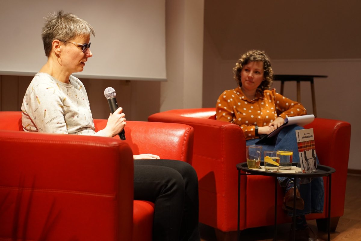 Dwie osoby siedzą naprzeciw siebie w jasnym pomieszczeniu na masywnych, czerwonych fotelach, biorąc udział w rozmowie. Osoba po lewej, zwrócona bokiem do aparatu, trzyma w dłoni mikrofon i ma krótkie, siwe włosy oraz jasną bluzę; na pierwszym planie widać jej nogi w czarnych spodniach. Po prawej siedzi druga osoba w koszuli w białe kropki na pomarańczowym tle, z notatkami na kolanach, lekko odchylona w fotelu. Między fotelami stoi mały, okrągły stolik, na którym leżą dwie szklanki z napojami i plasterkami cytryny oraz stojąca okładką do aparatu książka. W tle widoczny jest fragment ekranu projekcyjnego i zarys wysokiego stołu, co sugeruje kameralne spotkanie autorskie lub dyskusję publiczną.