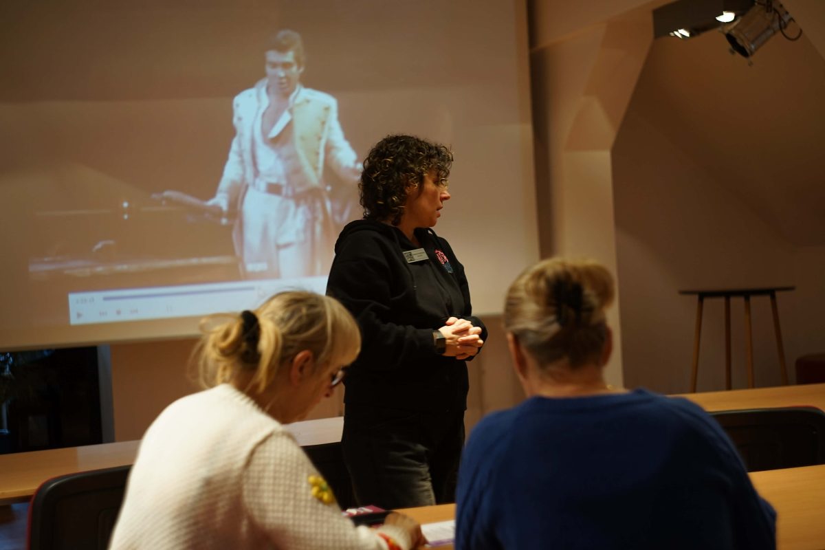 Kobieta stoi przy stole i prowadzi spotkanie lub wykład dla grupy osób siedzących w sali. Uczestnicy siedzą tyłem do aparatu i słuchają prowadzącej. W tle na ekranie wyświetlany jest obraz mężczyzny w jasnym stroju scenicz­nym, prawdopodobnie z nagrania wideo lub prezentacji. Sala ma charakter edukacyjny i spokojną, skupioną atmosferę.