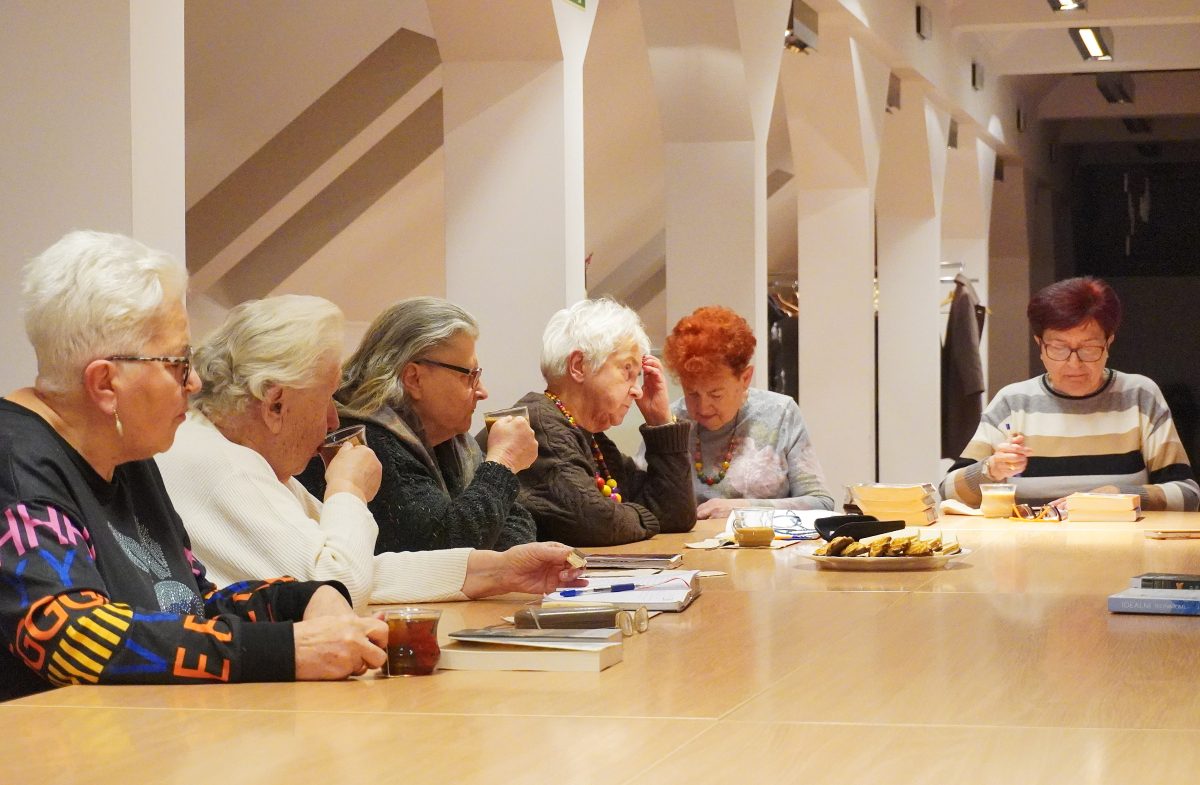 Grupa starszych kobiet siedzi przy długim, jasnym stole w nowoczesnym wnętrzu. Mają przed sobą książki, zeszyty i filiżanki z herbatą. Większość z nich czyta lub robi notatki, panuje spokojna, skupiona atmosfera wspólnego spotkania lub zajęć.