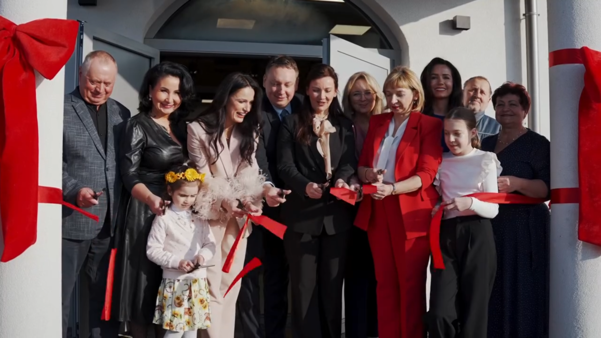 Grupa kilkunastu osób stoi ciasno przed wejściem do budynku podczas uroczystego przecięcia czerwonej wstęgi; dorośli i dzieci trzymają w dłoniach nożyczki oraz długie pasy czerwonej taśmy, a po bokach framugi drzwi przymocowane są wielkie dekoracyjne czerwone kokardy.