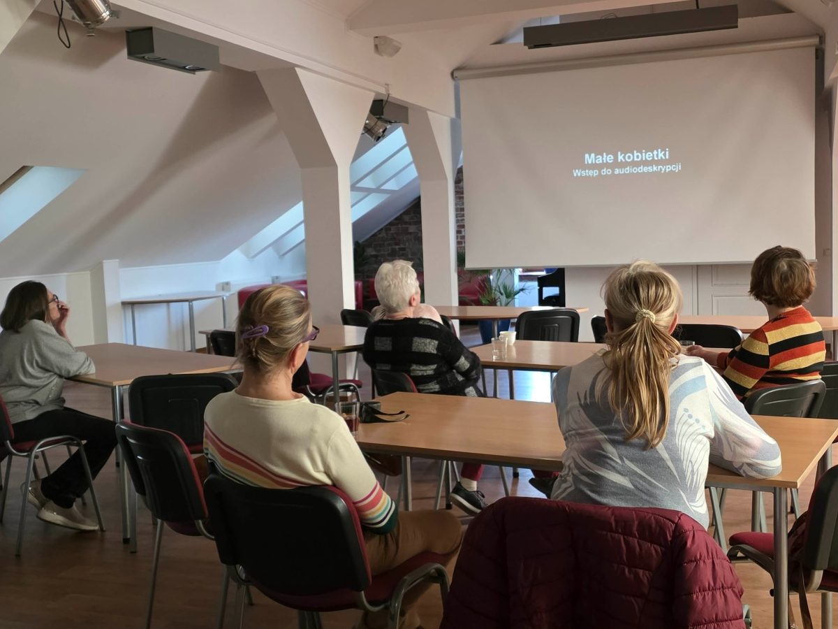Grupa kilku osób siedzi w luźno ustawionych rzędach tyłem do aparatu i ogląda ekran projekcyjny z napisem „Małe kobietki. Wstęp do audiodeskrypcji” na czarnym tle, w przestronnej, jasnej sali z białymi filarami i ukośnym dachem.