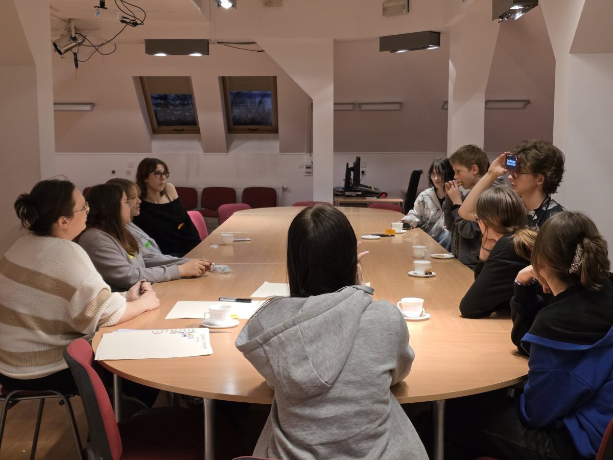 Grupa nastolatków i młodych dorosłych siedzi wokół długiego, owalnego stołu w jasnej sali spotkań na poddaszu. Po lewej stronie stołu siedzi kilka osób w swetrach i bluzach, patrzących w stronę prawej części stołu; przed nimi leżą kartki, długopisy i puste filiżanki po kawie. Po prawej stronie stołu siedzi reszta uczestników, część z nich trzyma w rękach kubki lub talerzyki z kawałkami ciasta. Jedna osoba po prawej, bliżej końca stołu, trzyma przy czole małe urządzenie z ekranem, jakby brała udział w grze lub ćwiczeniu. W tle widać skośny sufit z trzema oknami dachowymi, kilka punktowych lamp sufitowych i rząd pustych krzeseł przy ścianie.