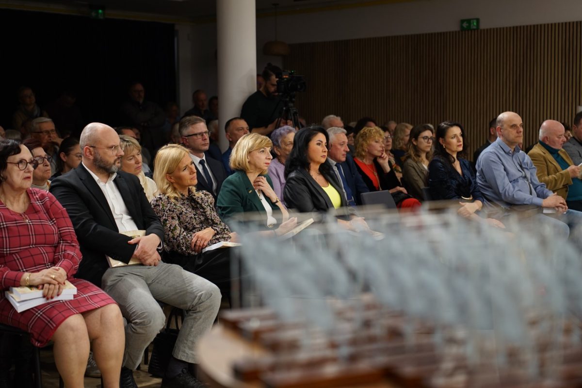 Grupa dorosłych osób siedzi w kilku rzędach na krzesłach w sali, skierowana twarzami w stronę niewidocznej sceny po lewej stronie kadru. W pierwszym rzędzie, na środku, siedzą elegancko ubrani mężczyźni i kobiety, część trzyma na kolanach książki lub notesy. W tle widać kolejne rzędy wypełnione publicznością oraz kamerzystę stojącego przy statywie, co sugeruje nagrywanie wydarzenia. Ściana po prawej stronie jest wyłożona pionowymi panelami, a przy suficie widać zielone tabliczki wyjścia ewakuacyjnego. Dolna część zdjęcia jest rozmyta i przedstawia rząd ustawionych obok siebie szklanych statuetek na stole przed publicznością.